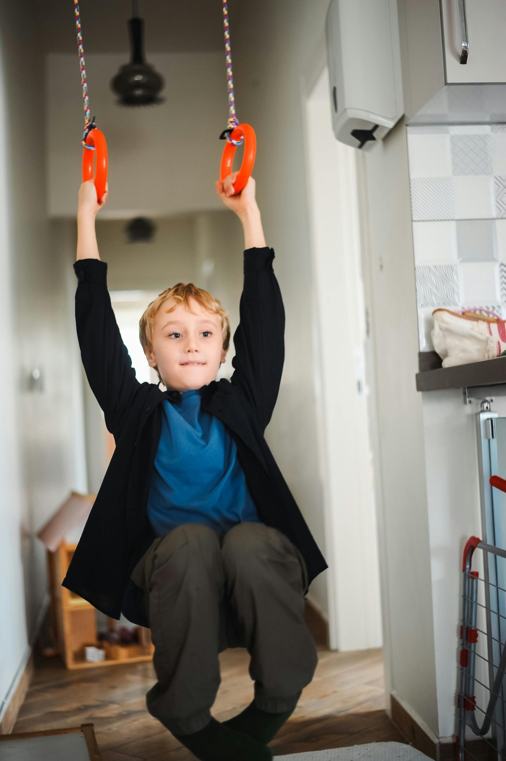 A young boy enjoying indoor play on bright orange gymnastic rings in a hallway.